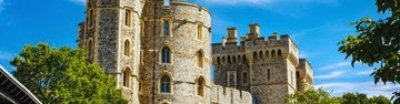 Stone castle towers under a bright blue sky with green trees in the foreground.