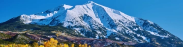 Snow-covered mountain rising above colourful woodland beneath a clear blue sky.
