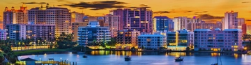 Waterfront city skyline with tall buildings reflected in calm water at sunset.
