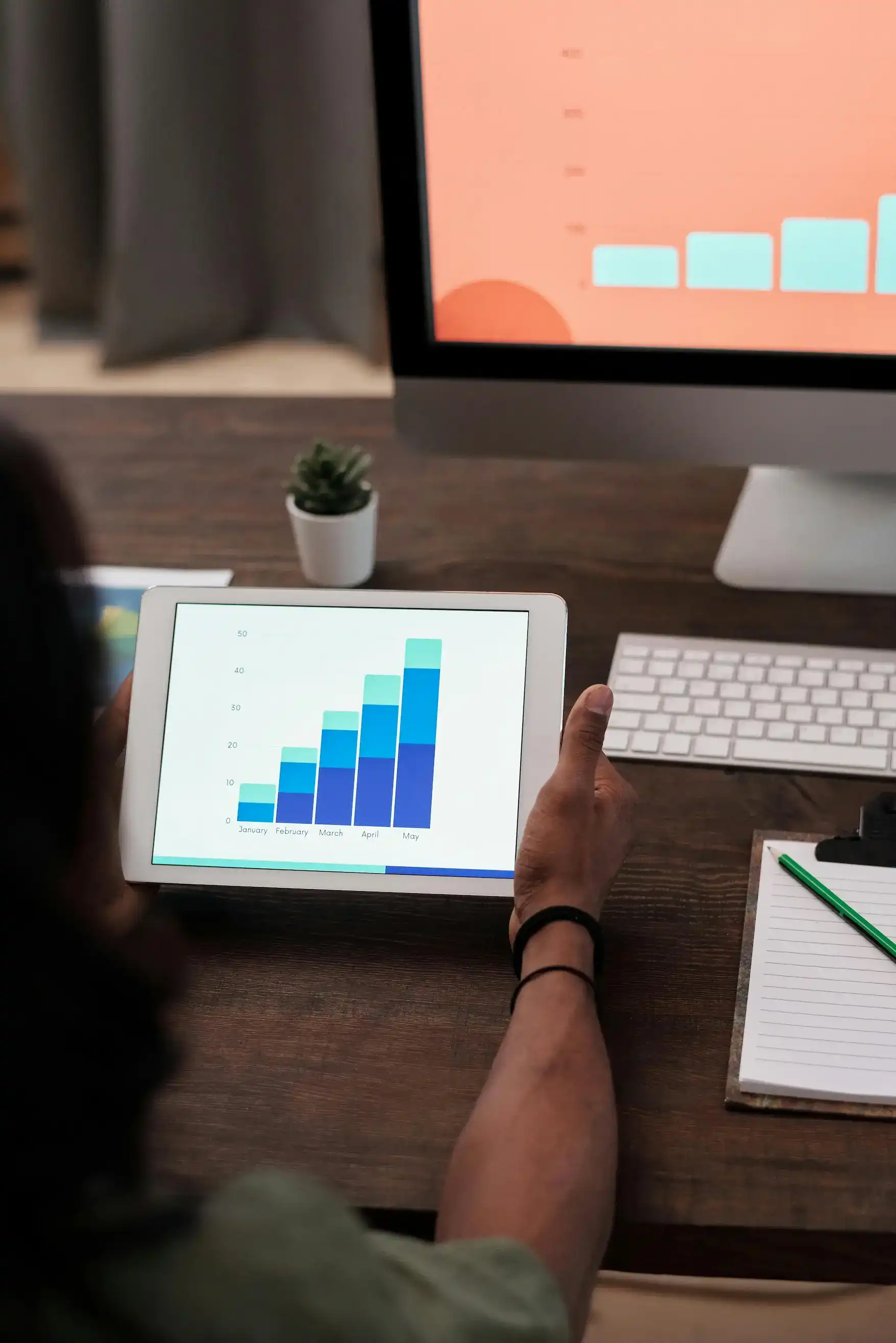 A person holding a tablet with a bar chart beside a desktop screen and notepad.